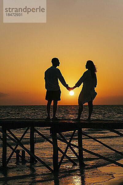 Silhouette eines Paares  das Händchen hält und bei Sonnenaufgang auf dem Pier steht