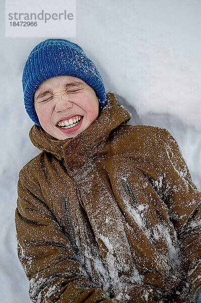 Fröhlicher Junge mit blauer Strickmütze liegt auf Schnee