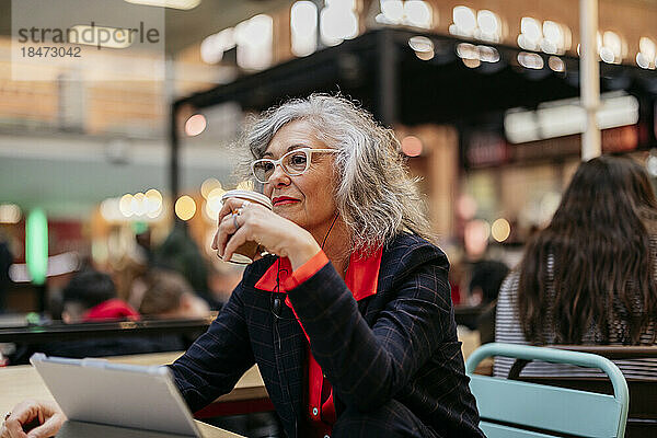 Nachdenkliche reife Geschäftsfrau sitzt mit Einwegkaffeetasse im Café