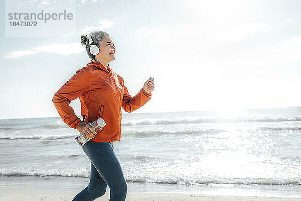 Lächelnde reife Frau mit Wasserflasche joggt an einem sonnigen Tag am Strand