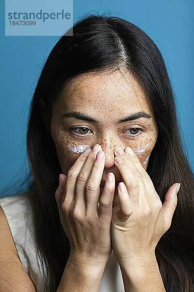 Woman with freckles applying lotion on face against blue background
