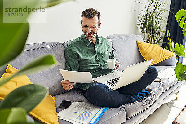 Glücklicher Mann  der von zu Hause aus arbeitet und sich Dokumente ansieht  während er mit einem Laptop auf dem Sofa sitzt und Kaffee trinkt