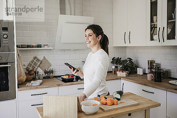 Glückliche Frau steht zu Hause mit Smartphone in der Küche