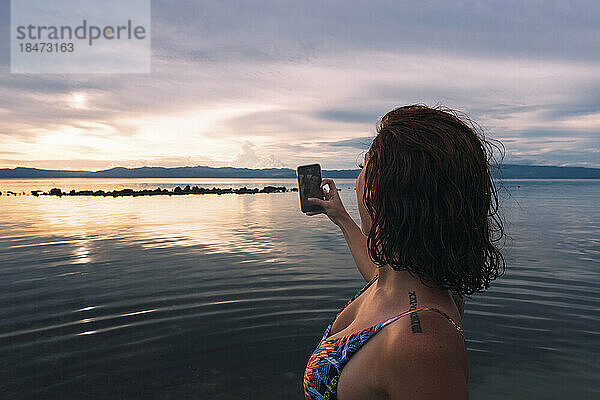 Junge Frau fotografiert Sonnenuntergang mit Smartphone am Strand