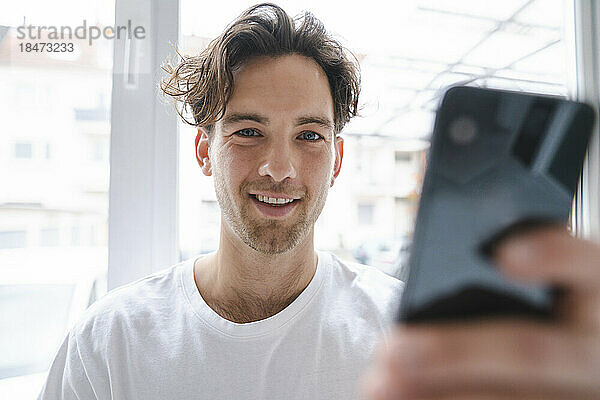 Lächelnder junger Mann mit Mobiltelefon vor dem Fenster