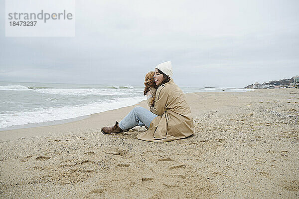Junge Frau sitzt mit Maltipoo-Hund im Sand am Strand