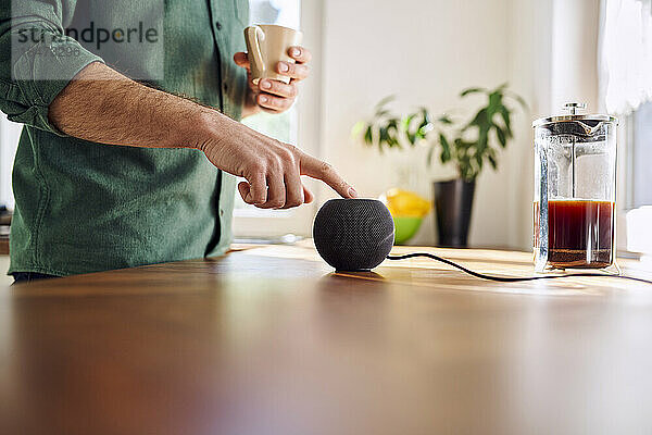 Mann steht zu Hause und hält Kaffeetasse in der Hand und schaltet Lautsprecher ein