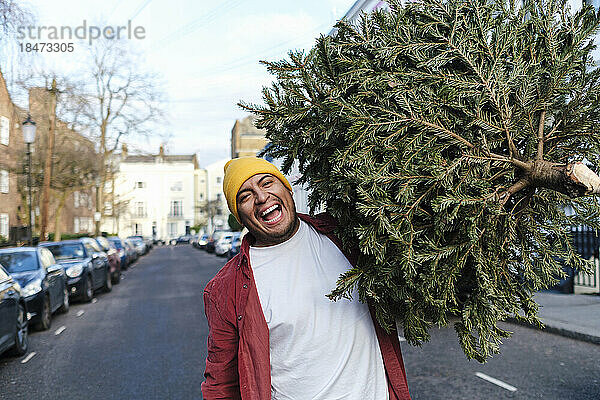 Fröhlicher Mann mit Weihnachtsbaum geht auf der Straße