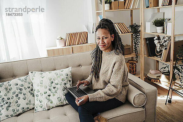 Lächelnde reife Frau mit Laptop sitzt zu Hause auf dem Sofa
