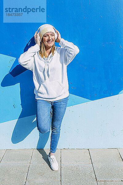 Glückliche Frau mit kabellosen Kopfhörern  die vor einer blauen Wand steht