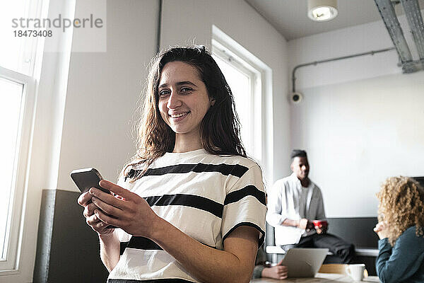 Glückliche Geschäftsfrau hält Smartphone mit Kollegen im Hintergrund