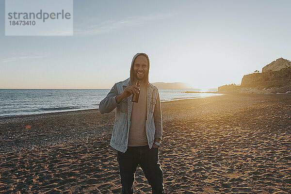 Glücklicher Mann steht mit einer Flasche Bier am Strand