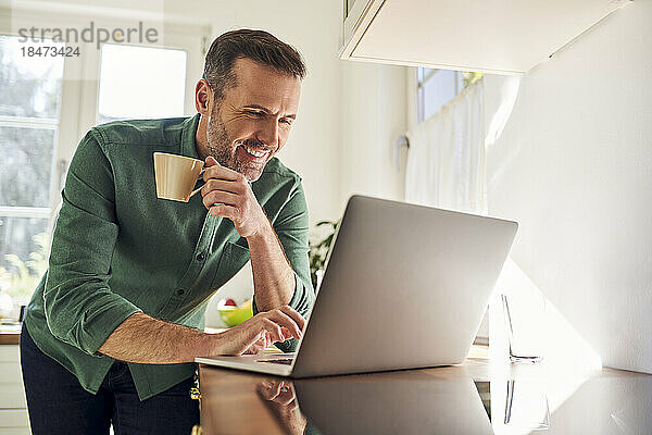 Smiling man using laptop while drinking coffee in the kitchen