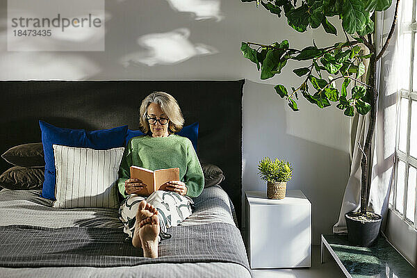 Reife Frau liest ein Buch und sitzt zu Hause im Bett