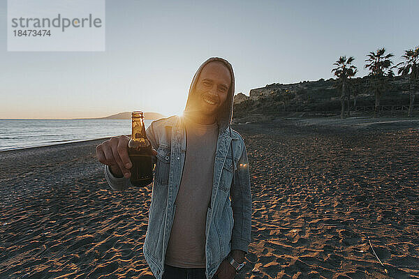 Lächelnder Mann steht bei Sonnenuntergang mit Bierflasche