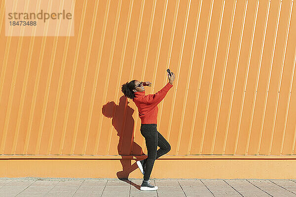 Frau macht Selfie mit Handy auf Fußweg vor orangefarbener Wand