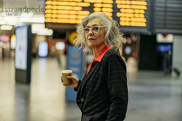 Reife Geschäftsfrau mit Einwegkaffeetasse steht am Bahnhof