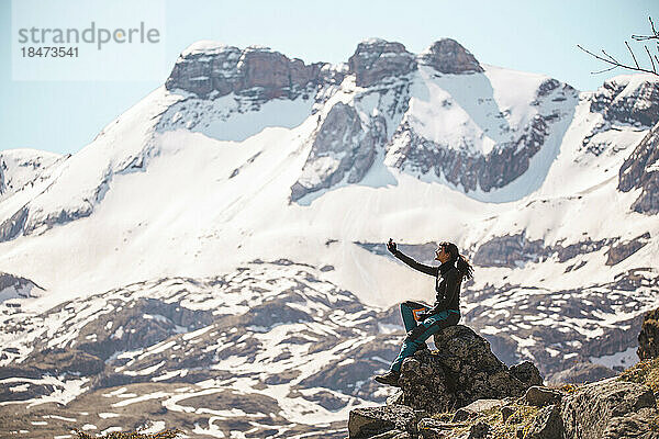 Junge Frau macht ein Selfie auf einem Felsen vor den Bergen