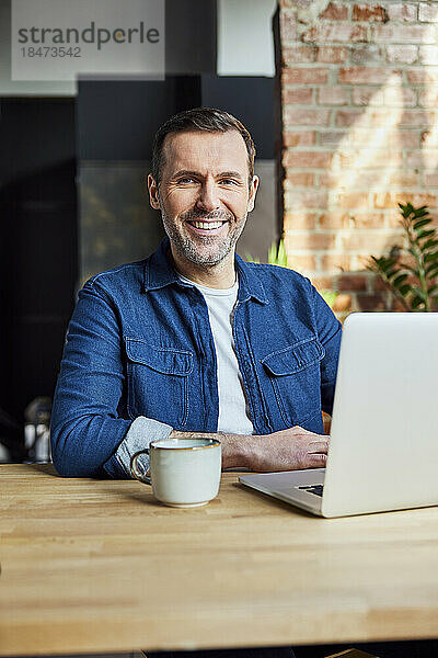 Lächelnder Freiberufler mit Laptop und Kaffeetasse am Schreibtisch im Loft-Büro