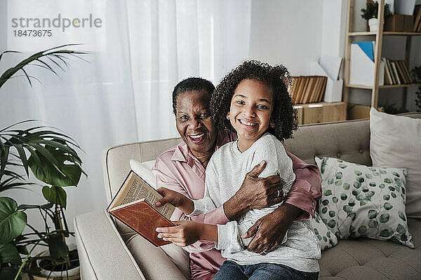 Glückliches Mädchen hält ein Buch in der Hand und sitzt mit Großmutter zu Hause auf dem Sofa