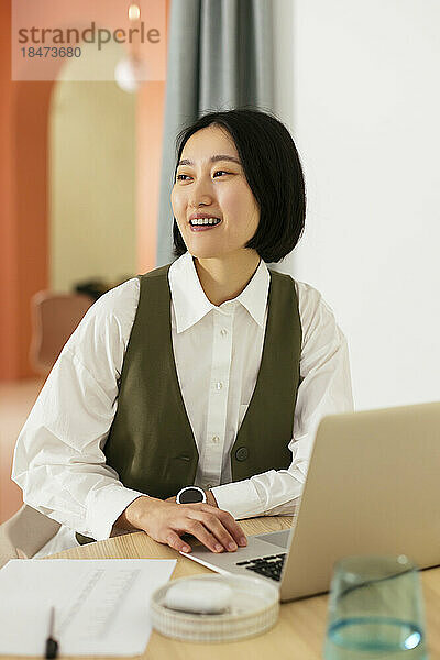 Glückliche Geschäftsfrau mit kurzen Haaren sitzt am Schreibtisch im Büro