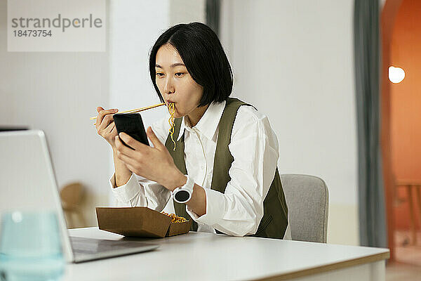 Geschäftsfrau isst Nudeln und benutzt Smartphone im Büro