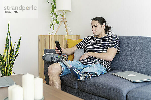 Junger Mann mit Beinprothese benutzt Smartphone auf Sofa zu Hause