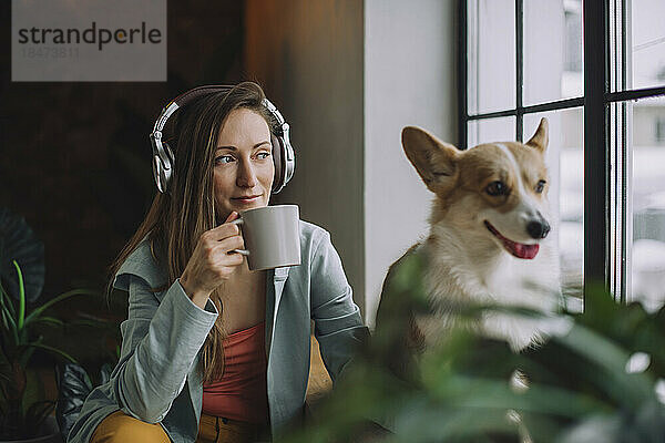 Frau trägt Kopfhörer und trinkt zu Hause Kaffee mit Hund