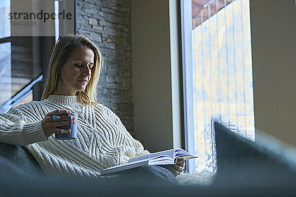 Frau liest Buch und trinkt zu Hause Kaffee