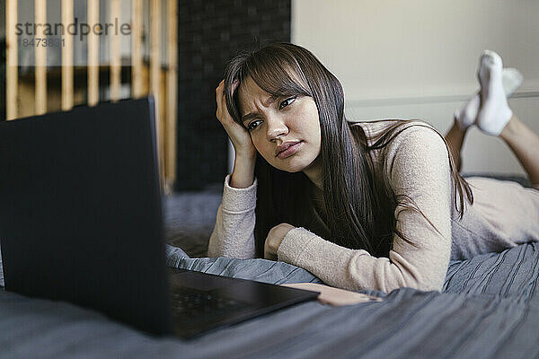 Junge Frau mit Kopf in der Hand und Laptop zu Hause im Bett