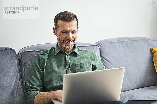Entspannter Mann  der von zu Hause aus mit einem Laptop auf dem Sofa arbeitet