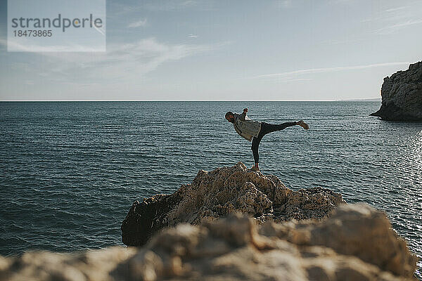 Mann balanciert auf einem Bein vor dem Meer an einem Felsen