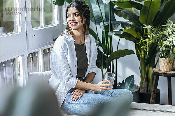 Glückliche junge Frau sitzt mit einem Glas Wasser am Fenster