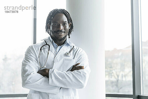 Lächelnder junger Arzt steht mit verschränkten Armen in der Klinik