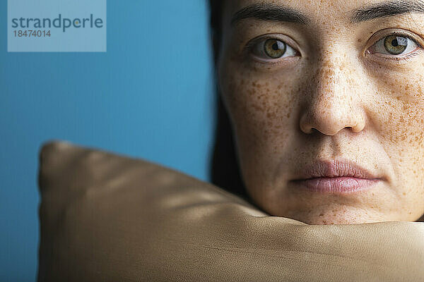 Woman with freckles resting chin on silk pillow against blue background