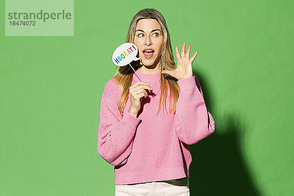 Woman gesturing and holding speech bubble with hurray text on it against green background