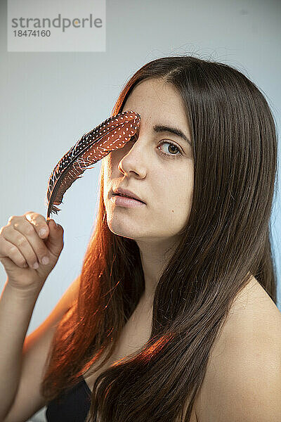 Young woman touching face with feather in front of wall