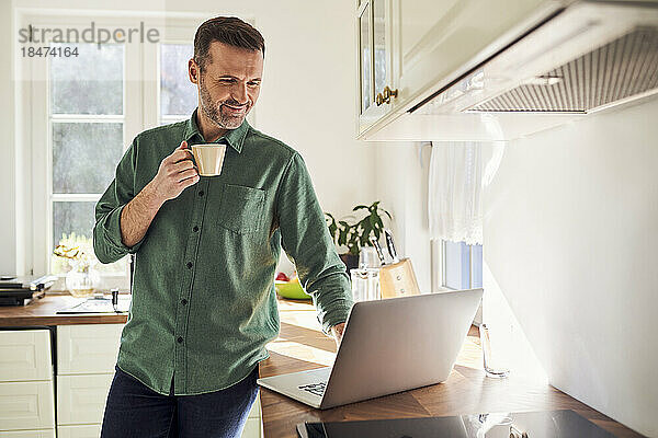 Lächelnder Mann benutzt Laptop  während er in der Küche Kaffee trinkt