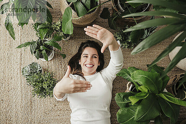 Glückliche Frau macht einen Fingerrahmen  der auf einem Teppich inmitten von Pflanzen liegt