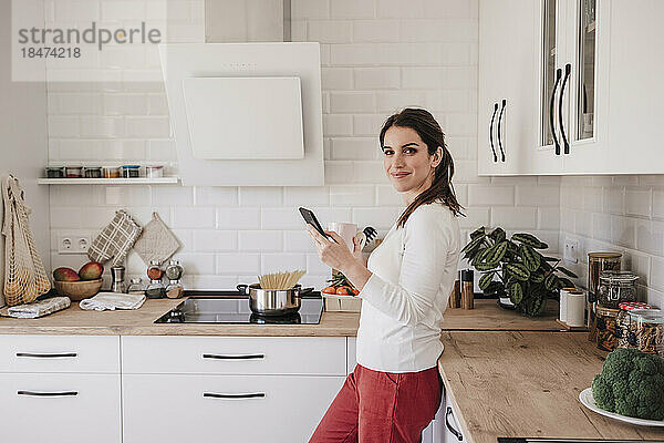 Lächelnde Frau mit Mobiltelefon steht am Tresen in der Küche