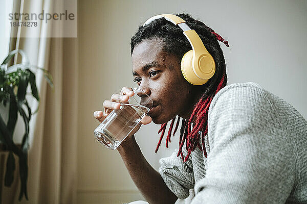 Nachdenklicher junger Mann mit Kopfhörern trinkt zu Hause Wasser