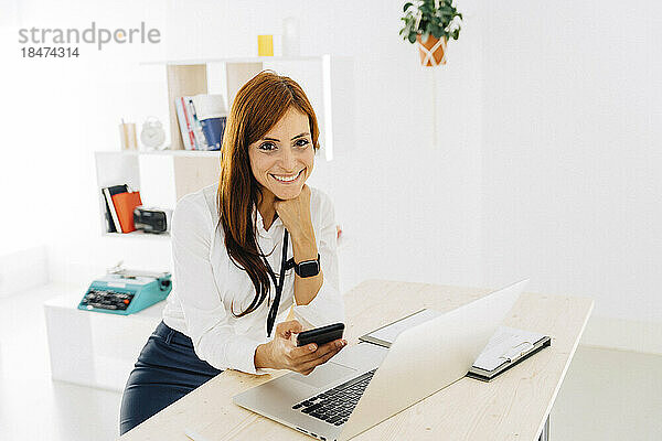 Lächelnde Freiberuflerin mit Mobiltelefon und Laptop am Schreibtisch