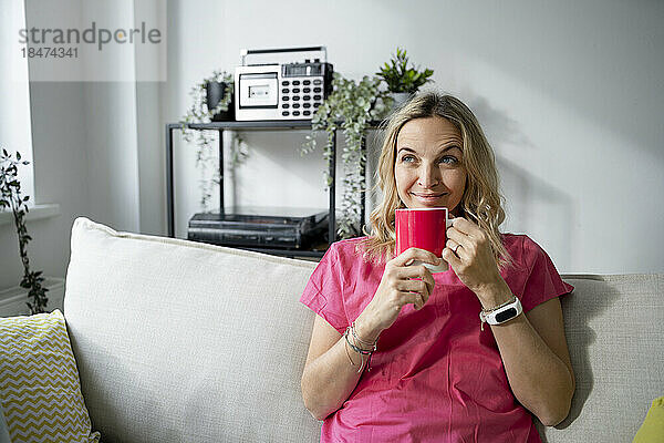 Lächelnde reife Frau mit Kaffeetasse sitzt zu Hause auf dem Sofa