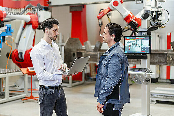 Kollege hält Laptop in der Hand und diskutiert mit Ingenieur in Roboterfabrik