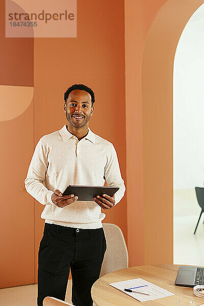 Lächelnder junger Geschäftsmann steht mit Tablet-PC im Büro