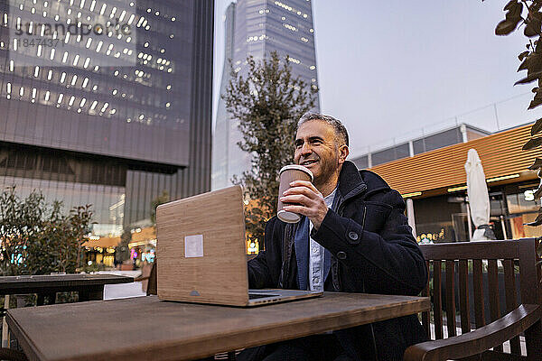 Glücklicher Geschäftsmann mit Kaffeetasse und Laptop am Tisch sitzend