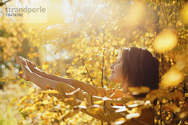Lächelnde Frau streckt die Arme inmitten goldenen Herbstlaubs aus