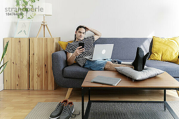 Junger Mann mit Beinprothese sitzt zu Hause auf dem Sofa und nutzt sein Smartphone