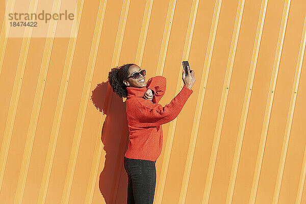 Glückliche Frau macht ein Selfie mit ihrem Smartphone vor einer orangefarbenen Wand