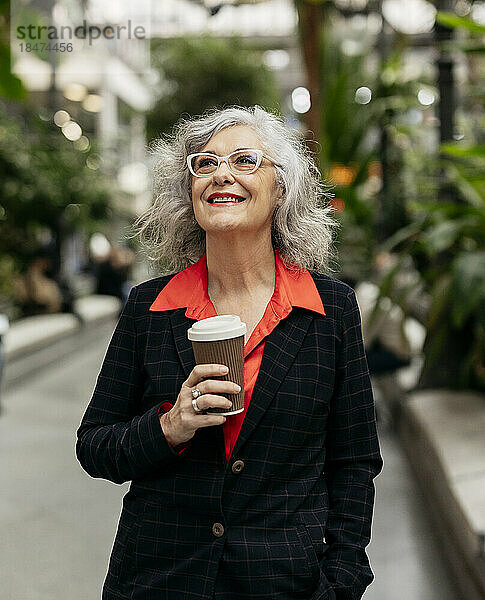 Nachdenklich lächelnde reife Geschäftsfrau mit Kaffeetasse steht im Garten